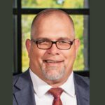 A middle-aged man with glasses, a goatee, and a bald head, wearing a suit, white shirt, and red patterned tie, smiling in front of a window with greenery outside.