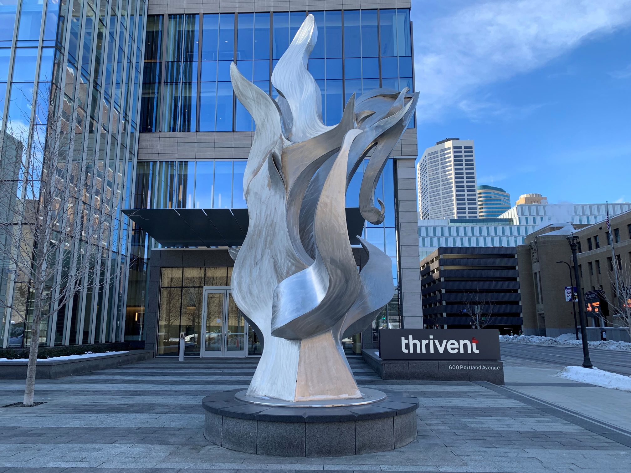 A large abstract silver sculpture stands outside a modern glass office building. The building has a thrivent sign near the entrance. Tall city buildings and a clear sky are visible in the background.