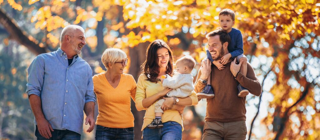 Three adults, a senior man and woman, a young couple, and two small children enjoy a walk outdoors in a park with autumn leaves, all smiling and interacting happily together.