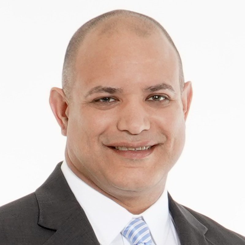 A man in a dark suit, white shirt, and light blue striped tie smiles at the camera against a plain white background.