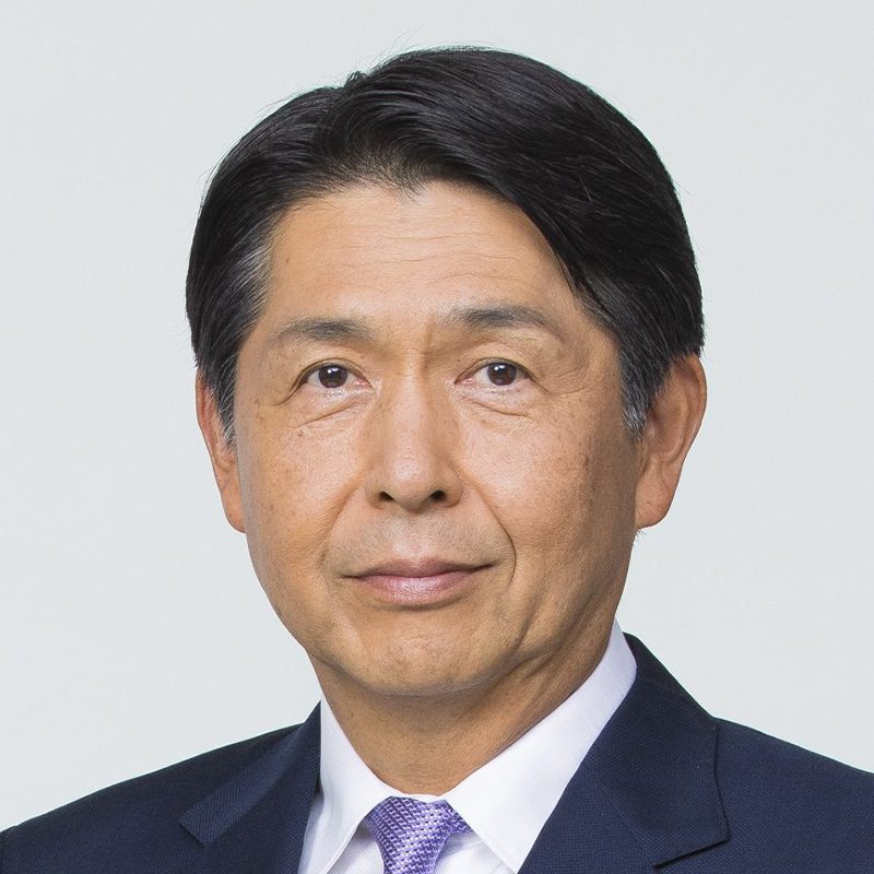 A middle-aged man with short, neatly combed black hair wearing a dark suit, white shirt, and light purple tie, posing against a plain light background.