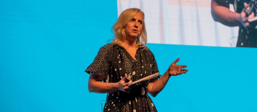 A woman with blonde hair in a black and white polka dot dress stands on stage holding a notebook, gesturing with one hand, in front of a bright blue background.