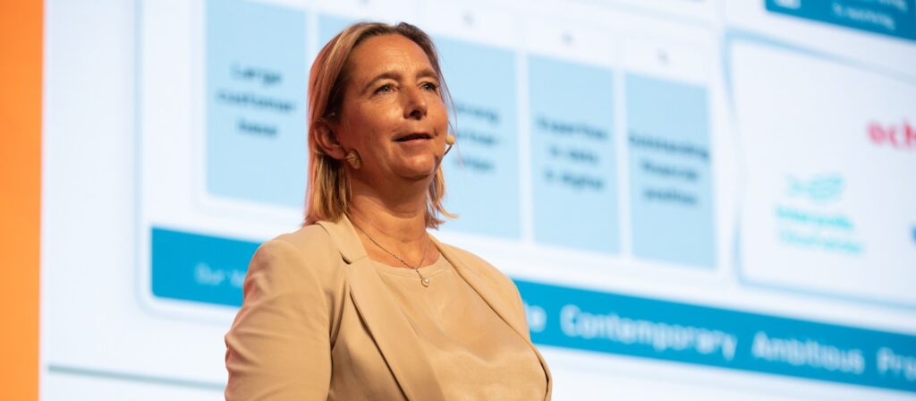 A woman in a beige blazer speaks on stage with a headset microphone. A blurred presentation slide with charts and text is projected behind her.