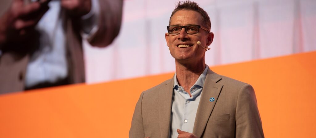 A man in a beige suit and glasses, wearing a headset microphone, smiles while speaking on stage in front of an orange and white background. His image is projected on a large screen behind him.