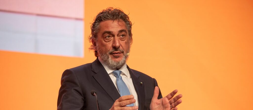 A man with curly gray hair and beard, wearing a dark suit, white shirt, and blue tie, speaks on stage in front of an orange background, gesturing with his hands. He has a headset microphone.