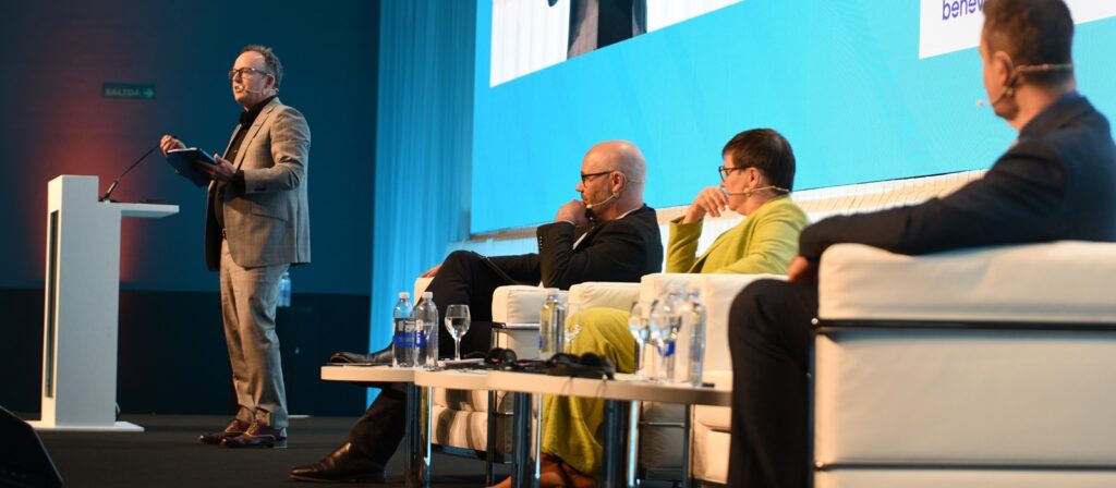 Four people participate in a panel discussion on stage; three are seated on white sofas, and one is standing at a podium speaking, with microphones and water bottles visible. A projection screen is in the background.