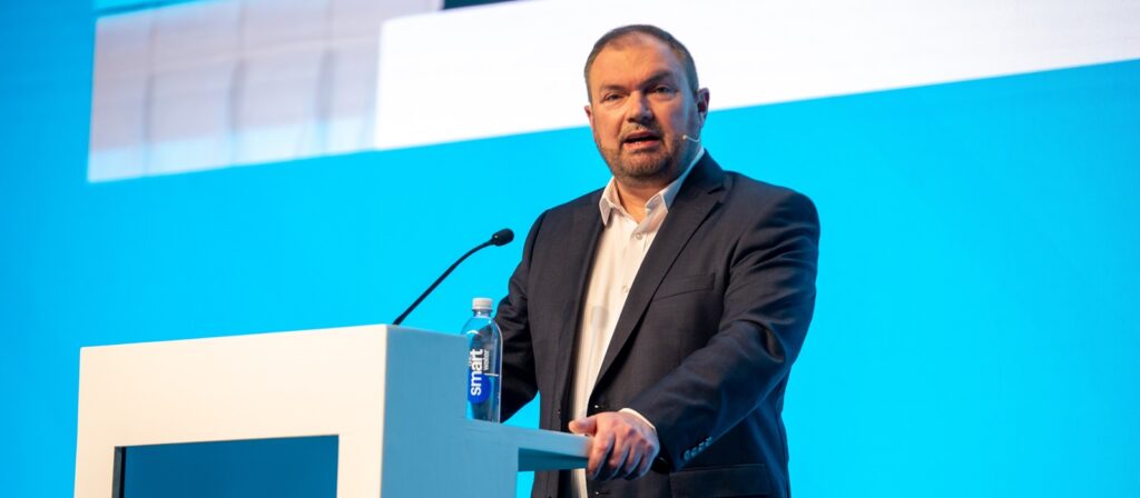 A man in a suit stands at a white podium with a microphone, speaking against a bright blue background. A bottle of water is on the podium.