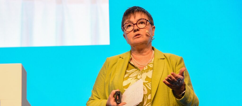 A person with short dark hair, glasses, and a green blazer speaks on stage with a headset microphone, holding papers in one hand and a device in the other, against a bright blue background.