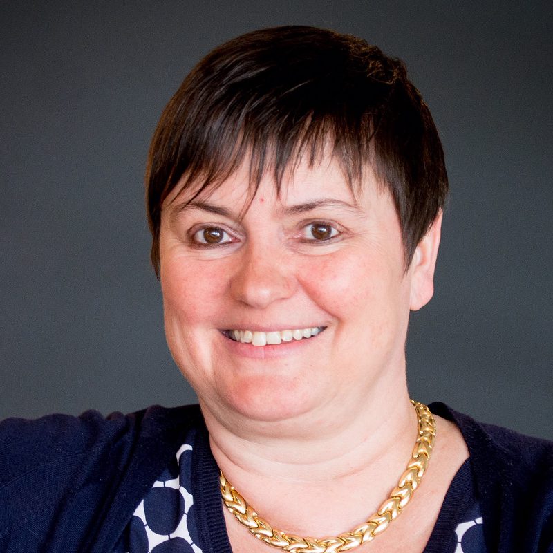 A woman with short dark hair smiles at the camera. She is wearing a gold chain necklace, a dark sweater, and a black-and-white patterned top, with a dark gray background behind her.