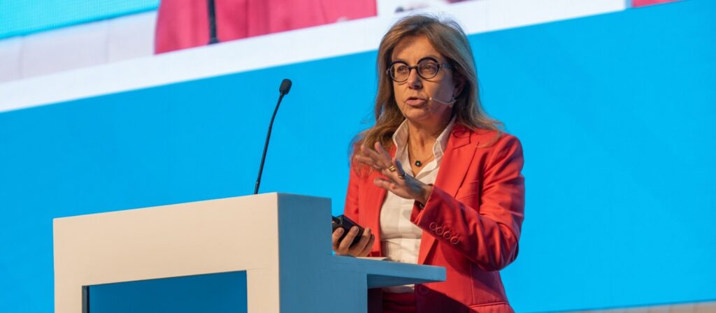 A woman in a bright red suit speaks at a podium with a microphone headset, gesturing with her hand. A blue and white background is visible behind her.