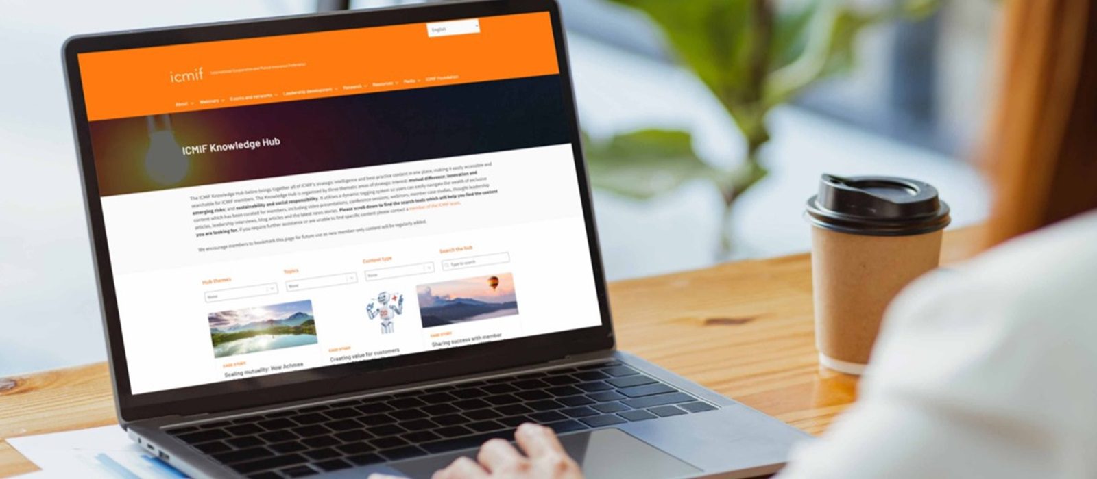 Person using a laptop on a wooden desk with a takeaway coffee cup nearby. The laptop screen displays the ICMM Knowledge Hub website, featuring an orange header and various categories.
