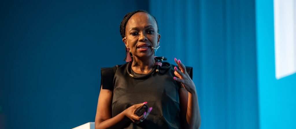 A woman wearing a black top and headset microphone speaks on stage, gesturing with one hand, in front of a blue background.