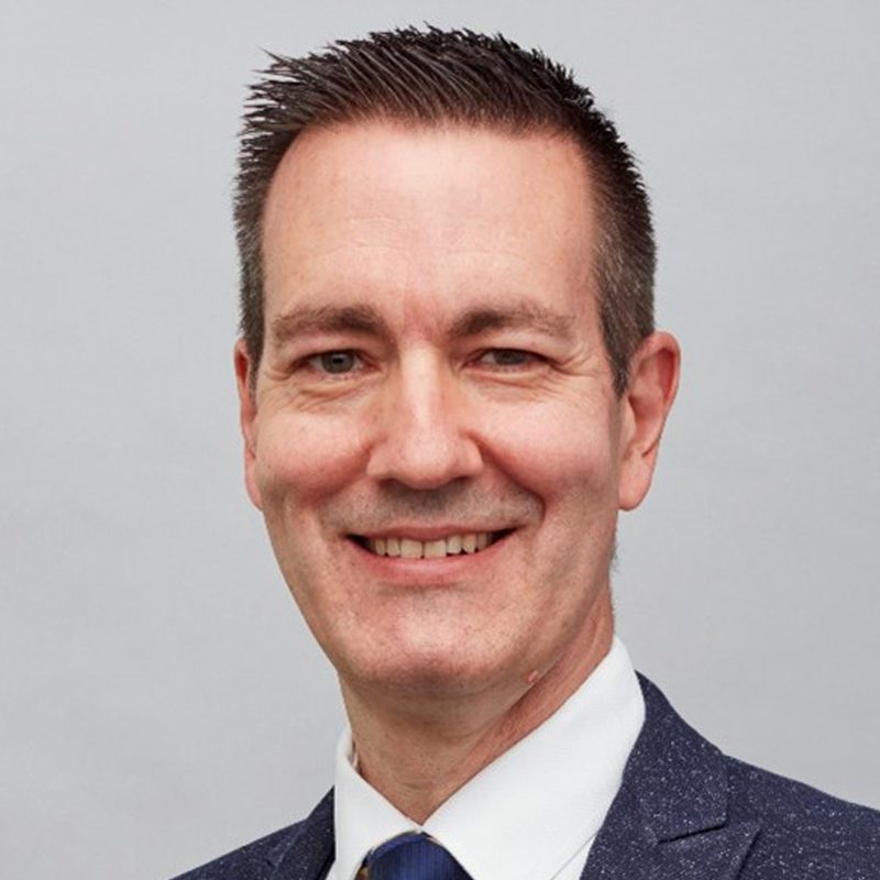 A man in a navy blue suit, white shirt, and blue tie smiles at the camera against a plain light gray background.