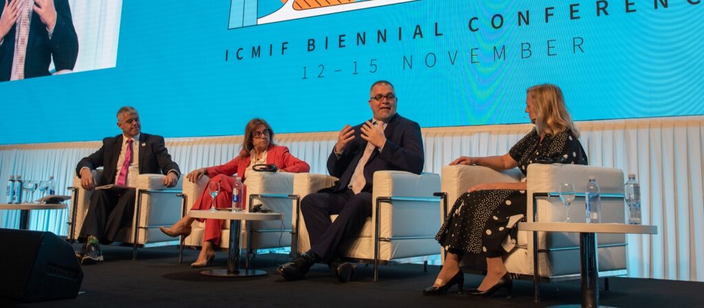 Four people in formal attire sit on a stage panel, speaking at the ICMIF Biennial Conference. A large blue screen behind them displays the event name and the dates 12-15 November. Bottles of water are on the table.