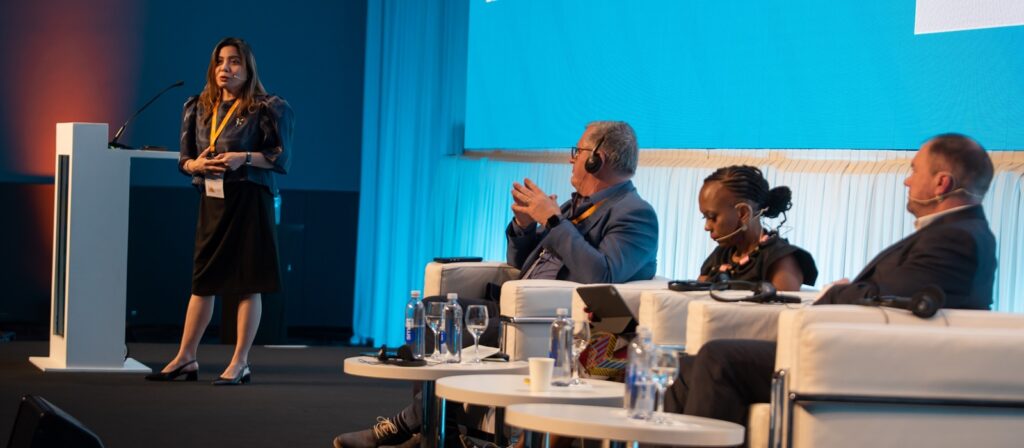 A woman stands and speaks at a podium on stage, while three seated panelists listen. All wear business attire and headsets, with a large blue screen in the background.