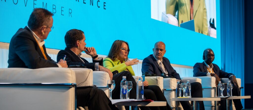 Five people in business attire sit on a stage panel, engaged in discussion. A woman in a yellow jacket speaks, while others listen. Water bottles and microphones are on the table. A large blue screen is behind them.
