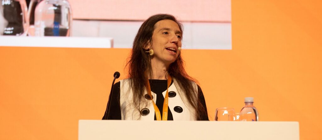 A woman with long brown hair speaks at a podium with a microphone, wearing a black and white outfit, against an orange backdrop. A glass of water and a bottle are on the podium.