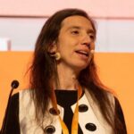 A woman with long brown hair speaks at a podium with a microphone, wearing a black and white outfit, against an orange backdrop. A glass of water and a bottle are on the podium.