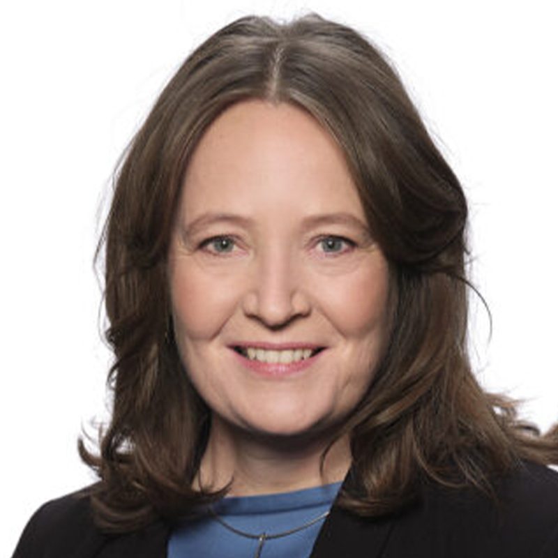 A woman with shoulder-length brown hair is smiling at the camera. She is wearing a blue top and a black jacket against a plain white background.