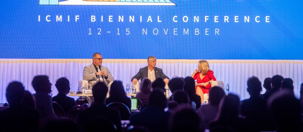 Three people sit on stage in front of an audience, speaking at the ICMIF Biennial Conference held from November 12-15. The stage background displays the event name and dates in large text.