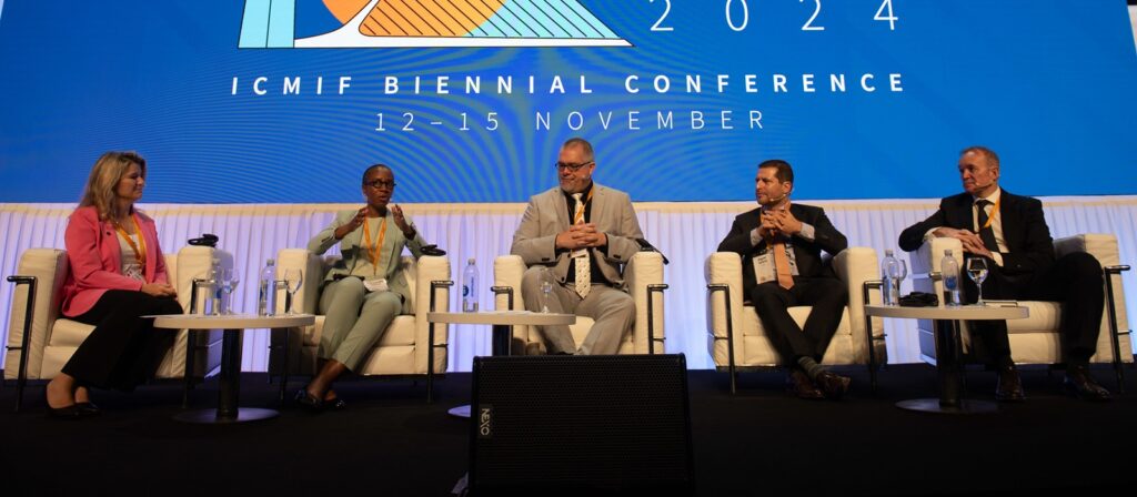 Five panelists sit on stage under a blue backdrop that reads ICMIF Biennial Conference 2024, 12-15 November. They are engaged in discussion, each with microphones, glasses of water, and conference lanyards.
