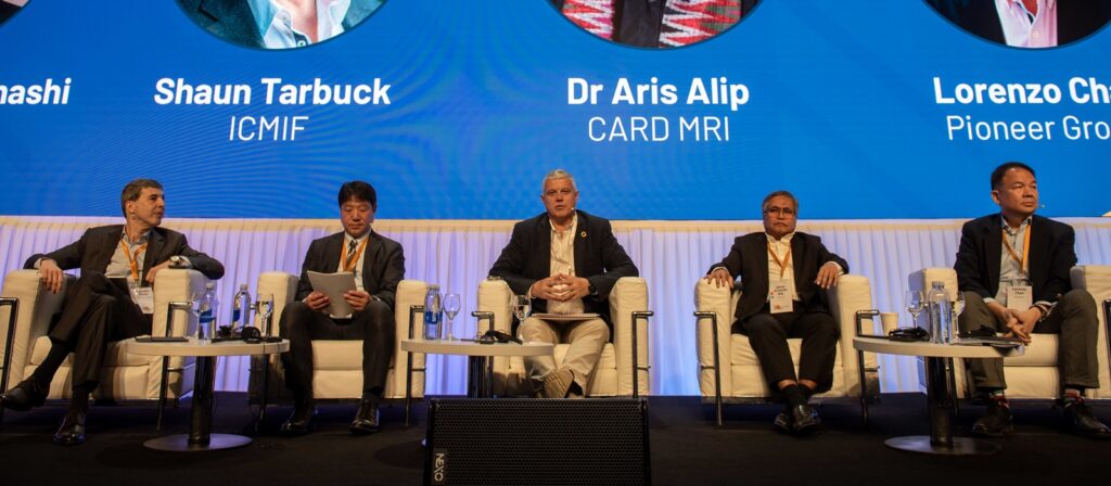 Five men in business attire sit on a stage in white chairs during a panel discussion. A blue screen behind them displays their names and organizations. Bottles of water and microphones are on tables in front of them.