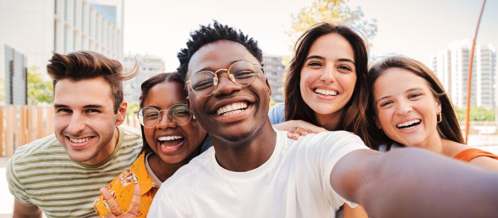 Five young adults stand close together outdoors, smiling and laughing at the camera as they take a group selfie. Buildings and trees are visible in the background on a sunny day.