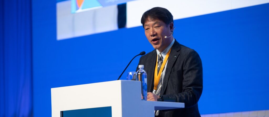 A man in a suit speaks at a white podium with microphones and a water bottle, wearing a headset microphone and yellow lanyard, against a blue background at a conference or event.