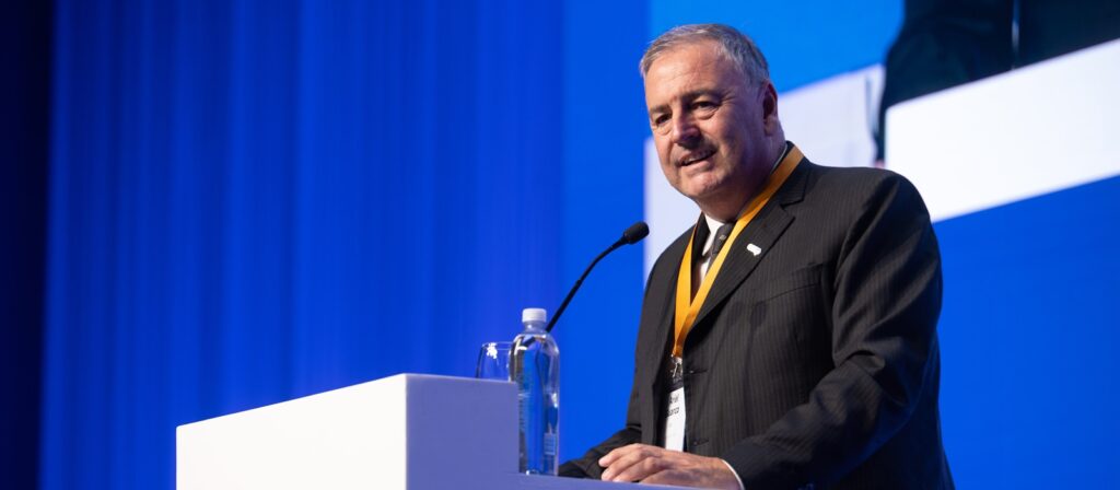 A man in a dark suit stands at a podium with a microphone, speaking at an event. He wears a yellow lanyard and a badge. A bottle of water and a glass are on the podium, with a blue background behind him.