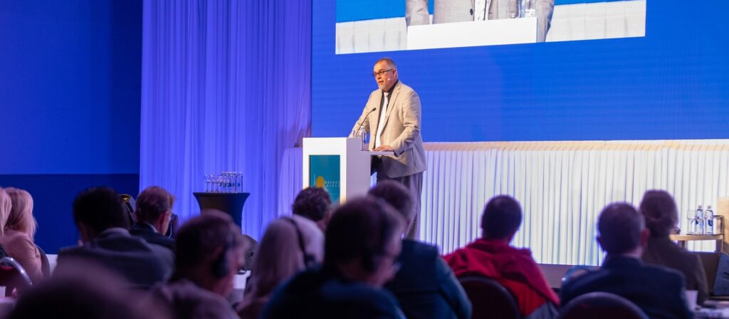 A man in a light suit speaks at a podium on a stage in front of an audience. A large screen behind him shows his image. The room is decorated in blue with white curtains and tables.