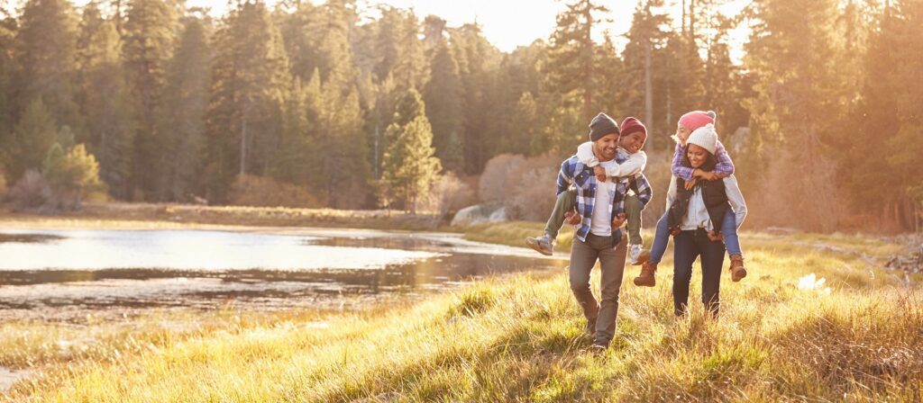 A family of four walks by a lakeside in a forest, with the adults giving piggyback rides to the children. They are dressed warmly and smiling, enjoying the outdoors on a sunny day.
