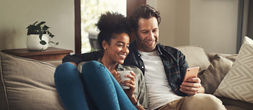 A smiling couple sits closely together on a beige sofa, looking at a smartphone. The woman holds a mug, and both appear relaxed and happy in a cozy, well-lit living room.