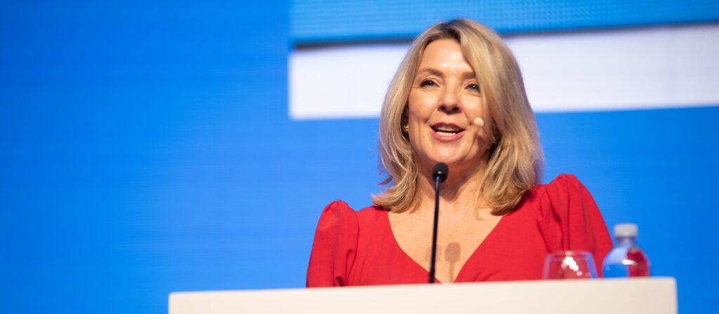 A woman with blonde hair, wearing a red dress, speaks at a podium with a microphone headset. The background is blue with a blurred graphic, and there is a glass and a bottle of water on the podium.