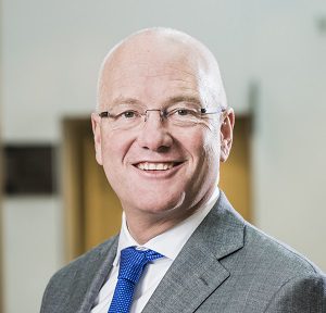 A smiling, bald man wearing glasses, a grey suit, white shirt, and blue tie stands in an indoor setting with a blurred background.