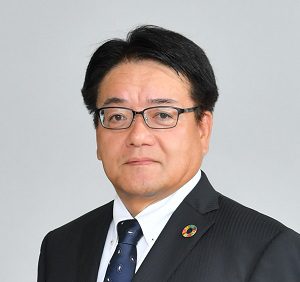 A man wearing glasses and a dark suit with a blue tie stands against a plain light gray background. He has short black hair and a circular pin with multiple colors on his lapel.