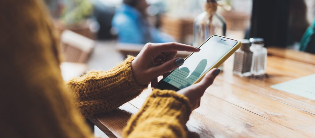 A person wearing a yellow sweater uses a smartphone at a wooden table in a cozy café, with blurred figures and soft light in the background.