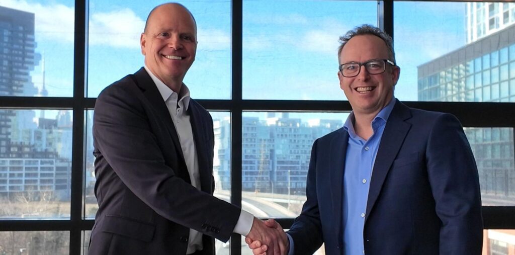 Two men in business suits smiling and shaking hands in front of large windows overlooking a cityscape with modern buildings.