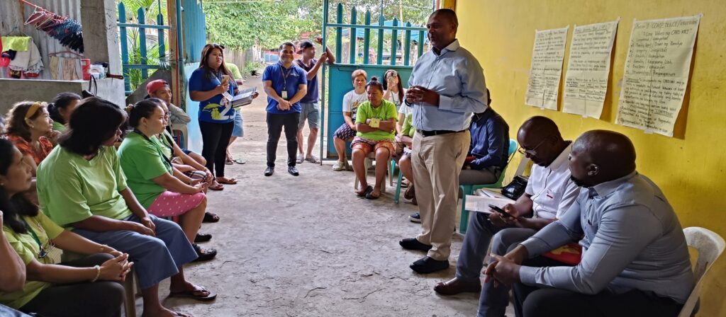 A man stands and speaks to a group of seated people inside a small room with yellow walls. Posters with handwritten notes are on the wall, and several people take notes while listening attentively.