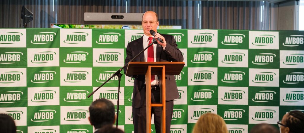 A man in a suit and red tie speaks at a podium with a microphone in front of an AVBOB-branded backdrop, addressing an audience.