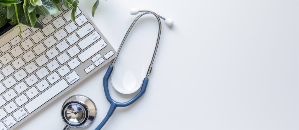 A stethoscope lying on a white desk next to a computer keyboard and a small potted plant, symbolizing healthcare and technology.