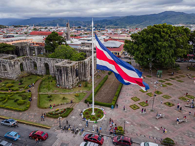 A large Costa Rican flag waves in a central plaza with people walking around; nearby are historic stone ruins, gardens, trees, and city buildings with mountains in the background under a cloudy sky.