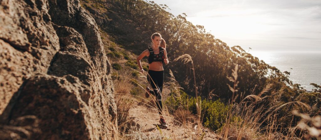 A woman in athletic wear runs on a narrow trail along a rocky hillside, with tall grass and trees around her. The sun is low in the sky, overlooking a scenic coastline in the background.