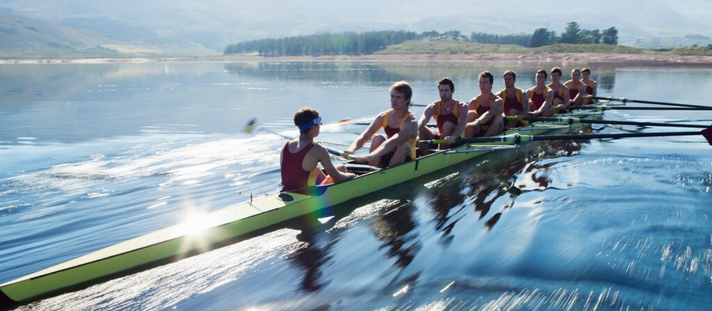 A team of eight rowers and a coxswain in a yellow rowing boat glides across a calm lake, surrounded by distant mountains and trees under a clear sky.