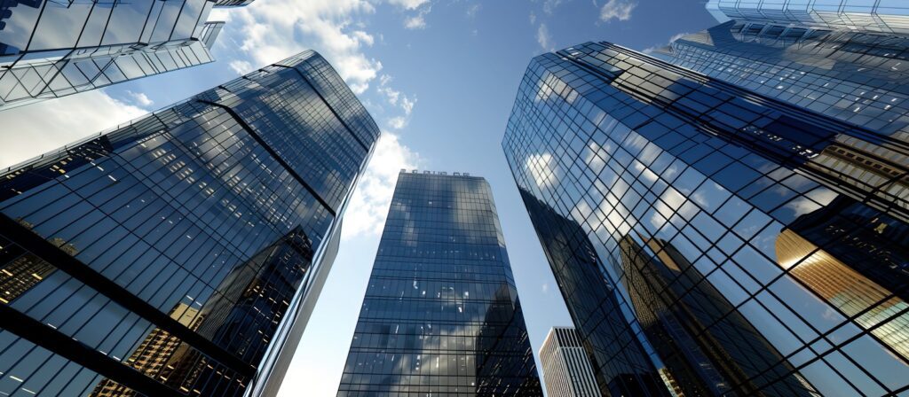 Tall glass skyscrapers reflecting the blue sky and clouds, viewed from below, with modern buildings surrounding them.