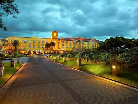 A brightly lit yellow hotel with a central tower and arches stands at the end of a paved driveway, surrounded by green lawns and bushes under a dramatic cloudy sky at dusk.