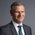 A middle-aged man with short gray hair, wearing a gray suit, white shirt, and blue striped tie, smiles slightly while posing against a plain gray background.