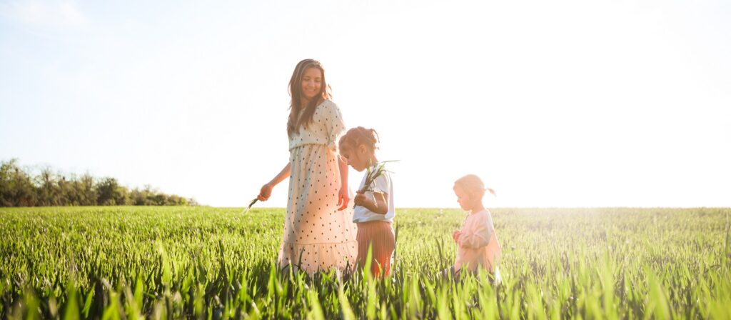 A woman in a white dress walks through a sunlit field with two young children. The children hold grass, and the three appear to be enjoying a peaceful moment outdoors surrounded by greenery.