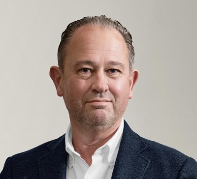 A middle-aged man with short, slicked-back hair and light facial stubble, wearing a white shirt and a dark blazer, poses against a plain, light background.