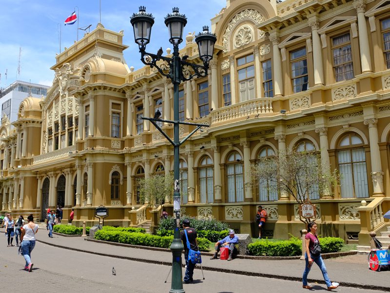 People walk and sit in front of a grand, ornate yellow building with arched windows and decorative details, under a blue sky. A lamppost stands in the foreground and a Costa Rican flag is visible on the roof.
