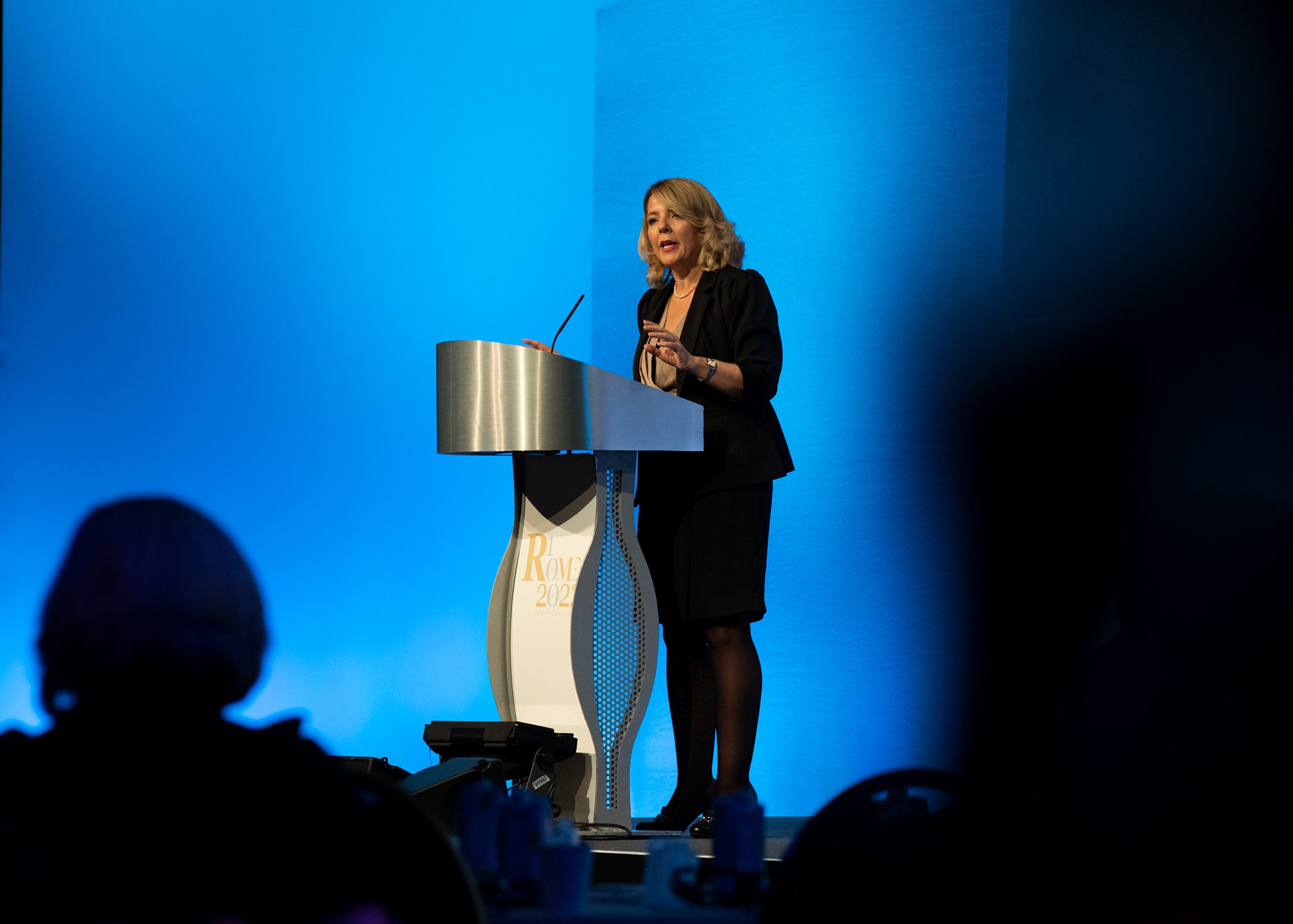 A woman in business attire stands and speaks at a podium on a stage with a blue background, addressing an audience at an event or conference.